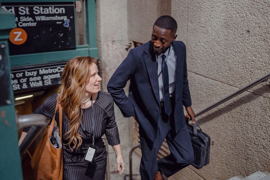 A man and a woman are ascending a staircase near West Drayton Station, engaged in a conversation. The woman has long, wavy red hair and is dressed in a dark pinstripe suit, carrying a brown leather sh