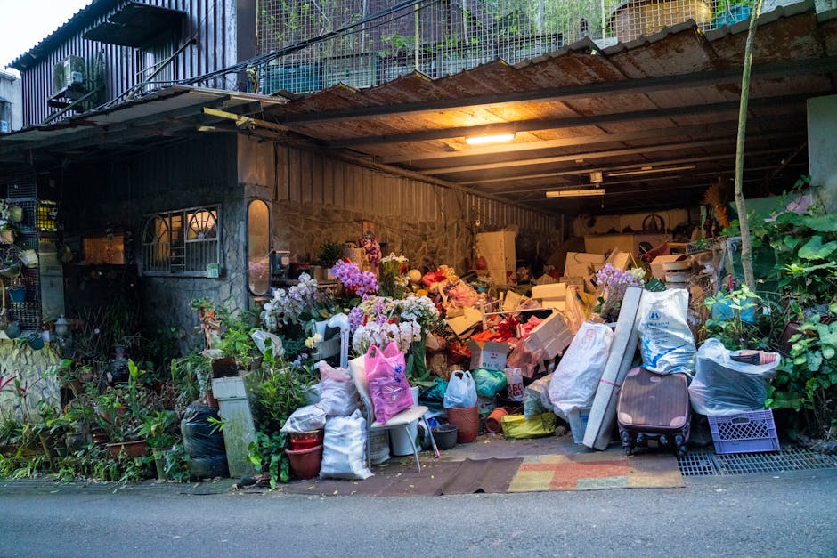 An outdoor storage area adjacent to a building, filled with various discarded items, including large bouquets of fresh flowers with pink, white, and purple blossoms, wrapped in paper and plastic. The 