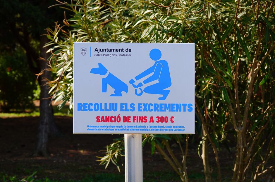 A rectangular signboard featuring a blue and white color scheme, placed outdoors among green leafy bushes. The sign displays a blue graphic of a person kneeling and picking up dog excrement with a sma