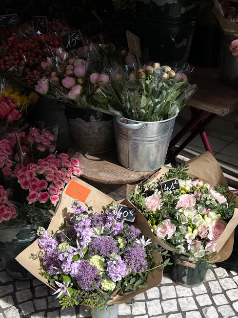 An arrangement of fresh flowers displayed outdoors on a wooden surface, featuring a bouquet of purple and lavender blooms with various green foliage wrapped in plain brown paper, alongside a smaller a